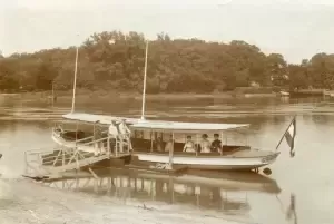 A vintage boat with several people docks at a wooden pier by a calm river surrounded by trees.