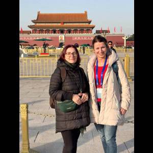 Two women stand in front of Tiananmen Square's iconic gate under clear skies, smiling for the photo.