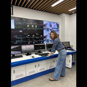 A woman stands at a control desk with monitors displaying train routes and surveillance in a modern facility.