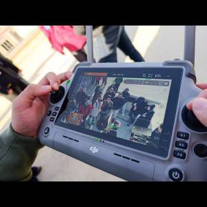 A person holds a DJI controller displaying a live view of a crowd on the screen.