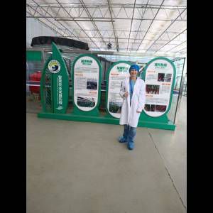 A person in a white lab coat and blue cap stands next to informational displays in a greenhouse setting.