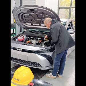 An older man inspects the engine of a silver car in a workshop, with a safety helmet visible in the foreground.