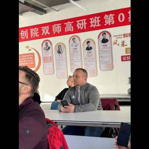 Students sit at tables in a classroom, with a prominent banner and profiles of teachers on the wall.
