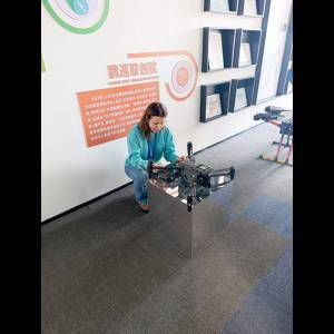 A woman inspects a drone displayed on a glass pedestal in a modern, well-lit room with framed certificates.
