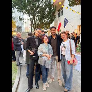 Four people pose together outdoors at an event, with trees and flags in the background, all smiling happily.