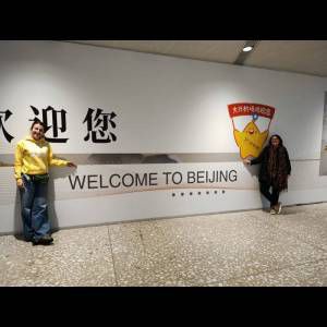 Two people pose by a wall that displays "WELCOME TO BEIJING" and a cheerful character illustration.