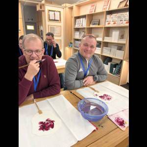 Two men enjoy a painting class, smiling with art supplies and a bowl of paint on the table.