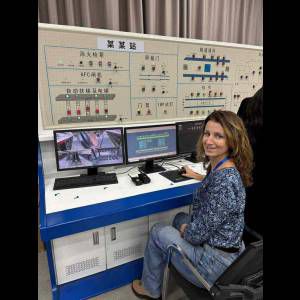 A woman sits at a control desk with monitors, overseeing a complex display of operational information.