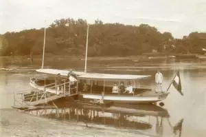 A historical boat with passengers docked by the shore, surrounded by trees and a calm river.