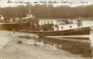 Historic photo of a boat named "RUDOLF" with people posing on the shore and water, surrounded by trees.