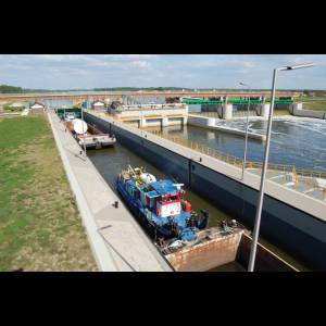 A blue tugboat is docked in a canal lock, surrounded by greenery and a dam, with cloudy skies above.