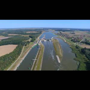 Aerial view of a river with a dam, surrounded by greenery and farmland, under a clear blue sky.