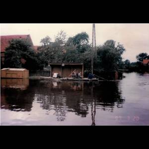 A group of people paddles a boat through a flooded area, with buildings partially submerged in the water. Rzeczyca/ Fot. J. Honc
