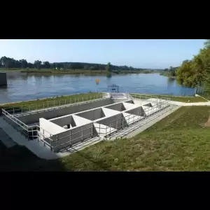 A concrete water treatment facility near a river, surrounded by greenery and a clear blue sky.