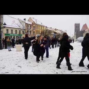 People are dancing in the snow-covered square, with colorful buildings and a tower in the background.