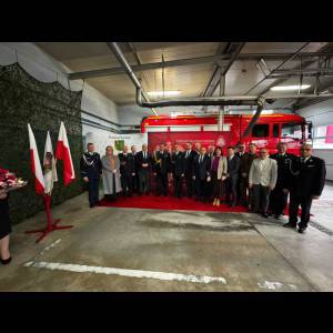 Group of people in formal attire pose with flags and a fire truck in a garage during a ceremony.