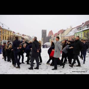 A group of people in formal attire playfully interacting in a snowy square, with colorful buildings and a tower in the background.