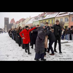 A group of people in winter clothing form a line in a snowy square, with colorful buildings in the background.
