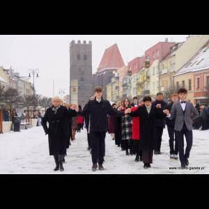 A group of people in winter attire hold hands, walking together in snow on a city street with historic buildings.