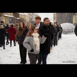 A group of people walks through a snowy street, dressed warmly, some chatting and wearing winter attire.