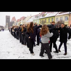 A line of people in winter clothing moves playfully on a snowy street, with colorful buildings in the background.
