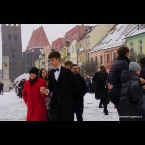 A group of people dressed formally walks on snowy ground near colorful buildings and a tower in winter.