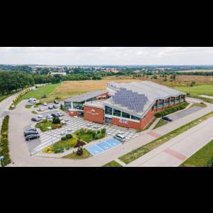 Aerial view of a modern building with solar panels, surrounded by grassy fields and parked cars.