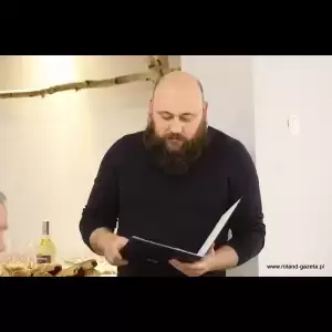 Klubu Honorowych Dawców Krwi „Kropelka Życia” A bearded man in a dark sweater reads from a document, surrounded by glasses and a bottle in a well-lit setting. Klubu Honorowych Dawców Krwi „Kropelka Życia”