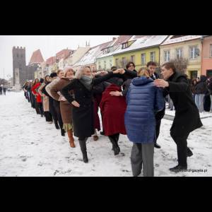 A line of people in winter clothing dances in the snow, with colorful buildings in the background.