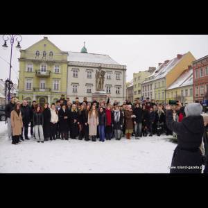 A large group of people gathers in the snow for a photo near a statue and colorful buildings.