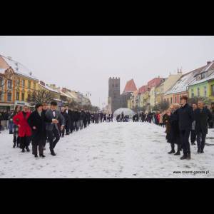 A snowy street scene with a line of people dressed up, surrounded by colorful buildings and a tower in the background.