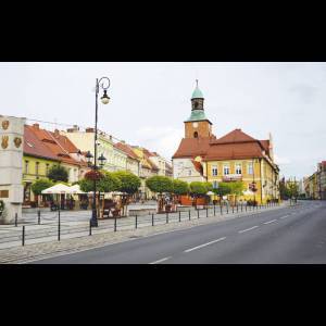 A quiet street scene featuring colorful buildings, a clock tower, and outdoor seating under lush greenery.