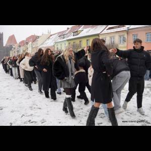 A line of people in winter clothes walking across a snowy street, with colorful buildings in the background.