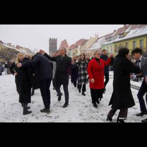A group of people in formal wear dance in a snowy square, with colorful buildings in the background.