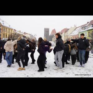 A group of people in winter clothing dancing on a snowy street, with colorful buildings and a tower in the background.