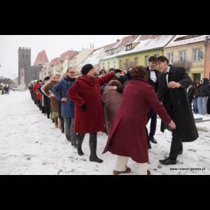 A long line of people in winter clothing gathers in a snowy square, engaging playfully amid colorful buildings.