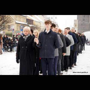 A group of people stands in a line on snowy ground, amid a town square with buildings in the background.