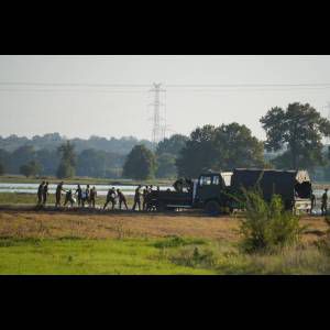Soldiers work in a field near a truck, with power lines and trees in the background under a clear sky.