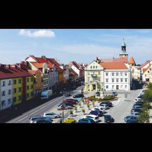 Colorful buildings line a bustling street with cars parked and a clock tower visible, under a clear blue sky.