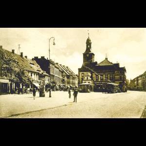 Historic town square with a clock tower, vintage streetcars, and people walking among traditional buildings.