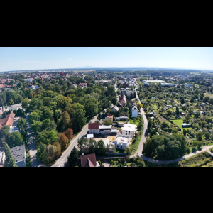 Aerial view of a residential area with forests, gardens, and distant hills under a clear blue sky.