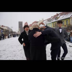 A snowy street scene featuring people interacting, dressed in winter attire and formal wear, with buildings in the background.