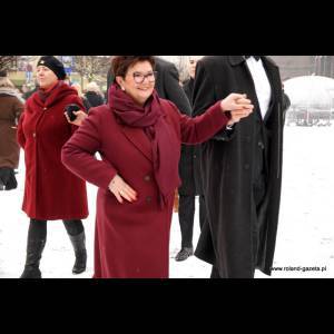 A smiling woman in a red coat and scarf poses in the snow, surrounded by elegantly dressed people.