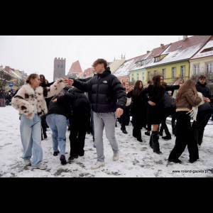A lively group of young people dance together in a snowy street, surrounded by colorful buildings.