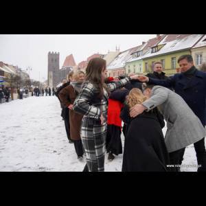 A snowy square filled with people in winter attire dancing and celebrating outdoors, with colorful buildings in the background.