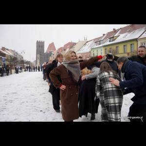 People are lined up in a snowy square, some interacting playfully while buildings and a tower are in the background.