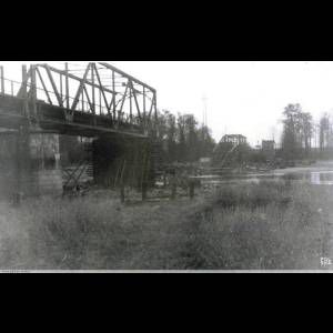 A dilapidated metal bridge over a river, surrounded by trees and tall grass, captured in black and white.