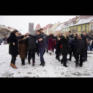 People are dancing in a snowy square, surrounded by colorful buildings and a historic tower in the background.