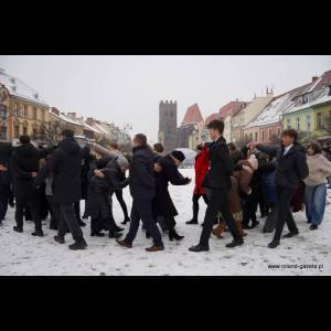 A group of people in winter clothes walking through snowy streets, with colorful buildings and a tower in the background.