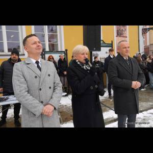 A group of people stand outside in winter attire, with three in front speaking at a public event.